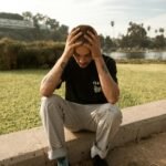 A young man sits on a ledge outdoors, appearing deep in thought and contemplation.